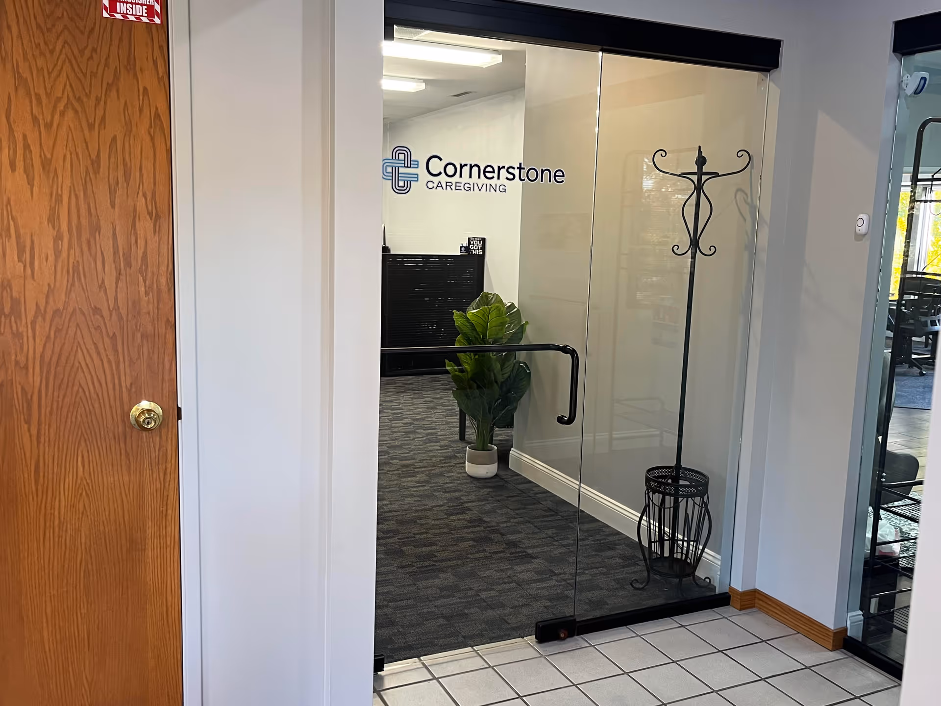 Glass doors with Cornerstone Caregiving logo open into a hallway with a coat rack and plant.