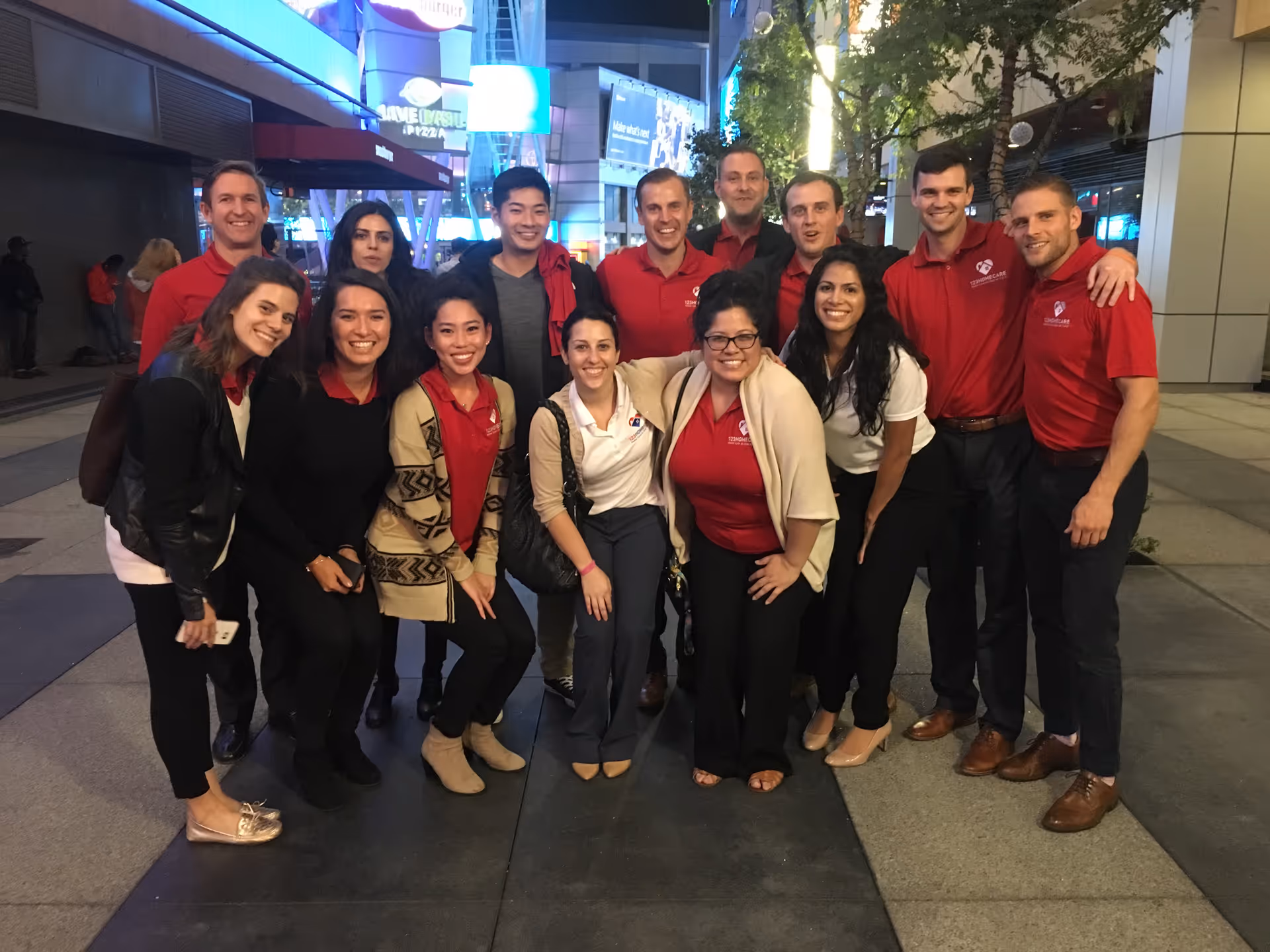 Group of staff in red shirts posing together on city sidewalk at night.