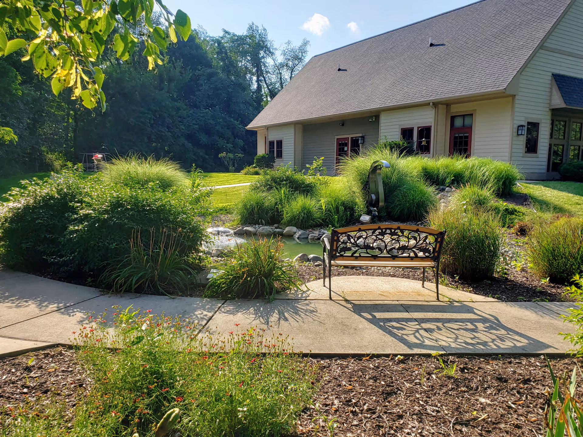 Outdoor garden area with bench, pond, and lush landscaping beside a light-colored senior facility.