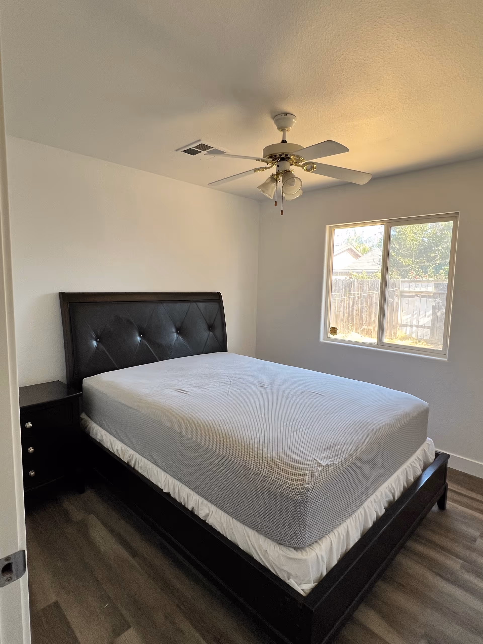 A simple furnished bedroom with a dark bed frame, nightstand, ceiling fan, and window.