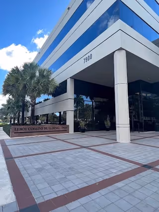 Modern glass-front building entrance with white pillars, palm trees, and exterior signage.