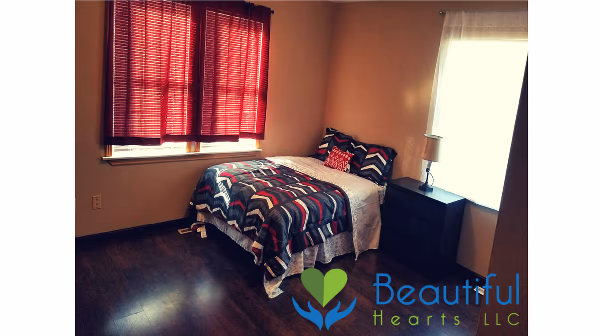 Bedroom with a bed, nightstand, lamp, red curtains, and Beautiful Hearts LLC logo.