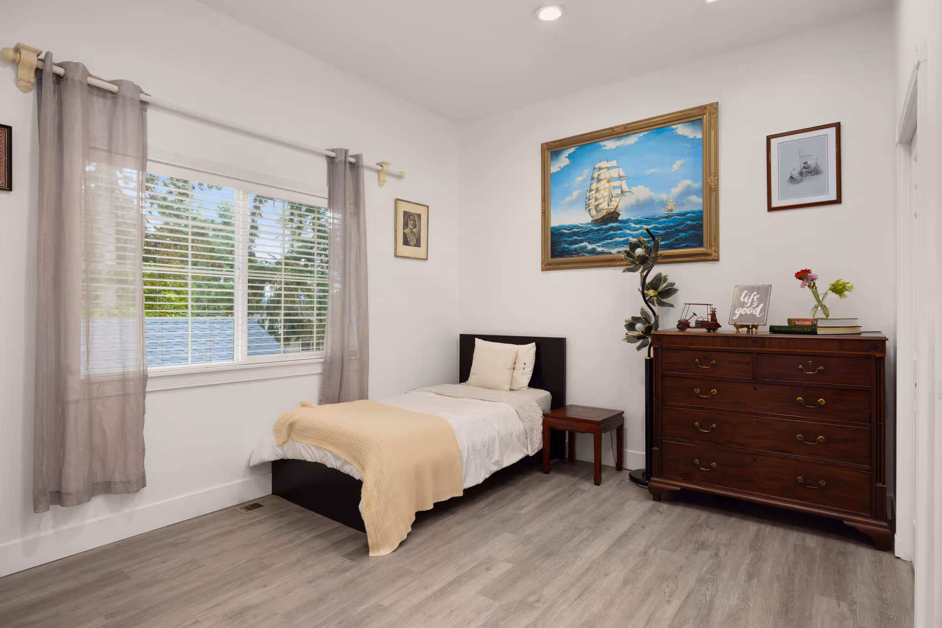 A cozy bedroom with a single bed, dresser, window with sheer curtains, and wall art.