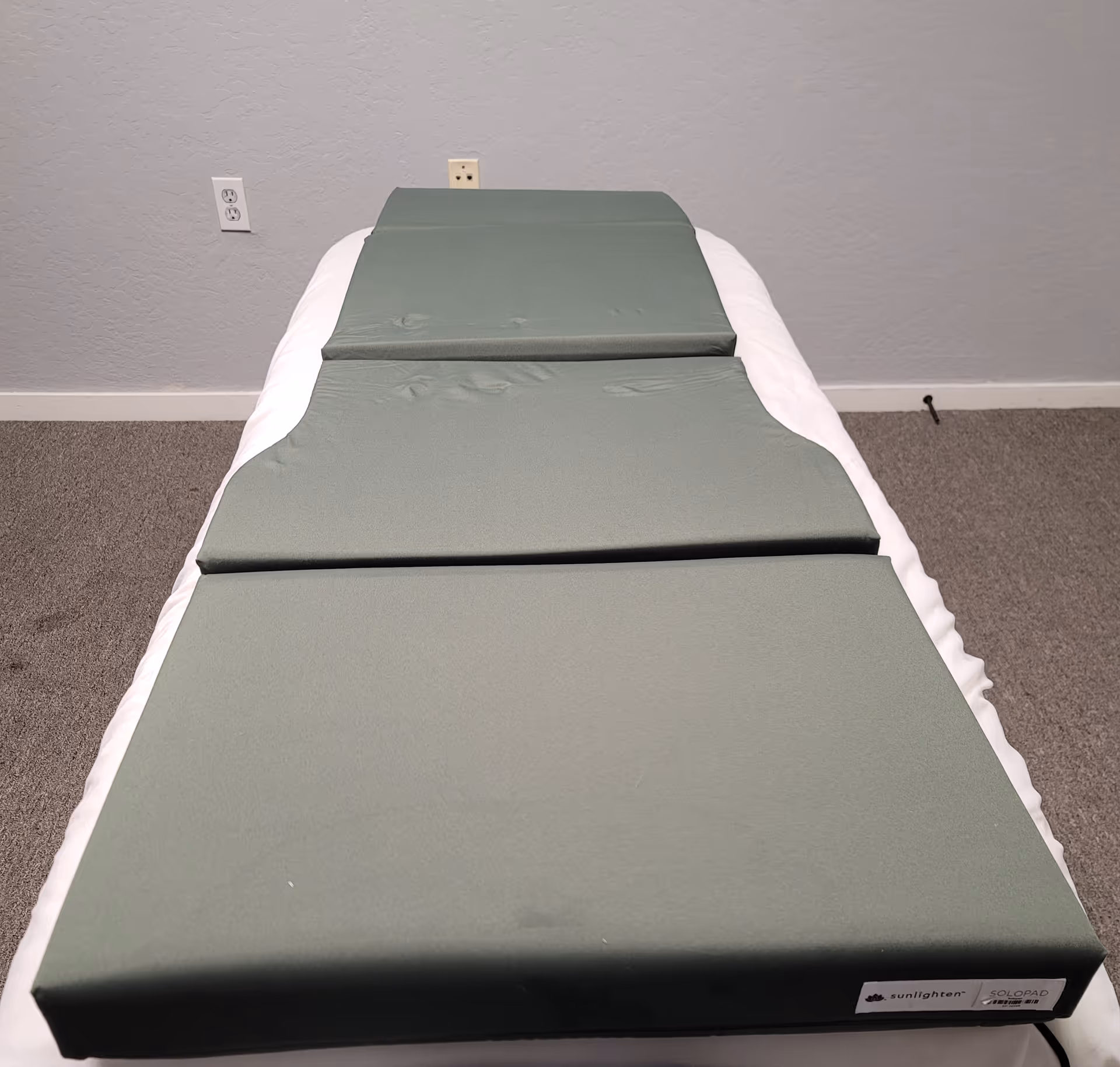 A padded massage table sits in a plain, carpeted room with gray walls.