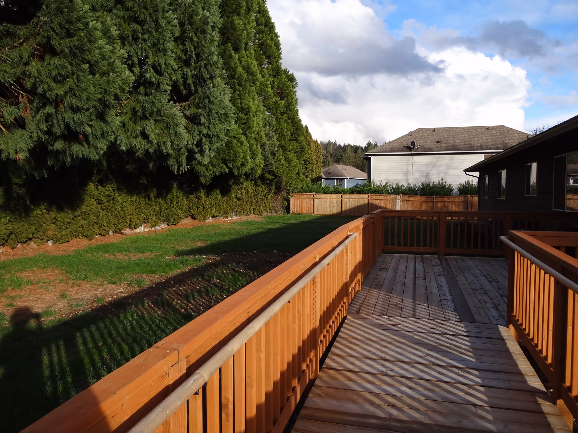 Wooden deck with railing extends from a house to a grassy backyard lined with trees.