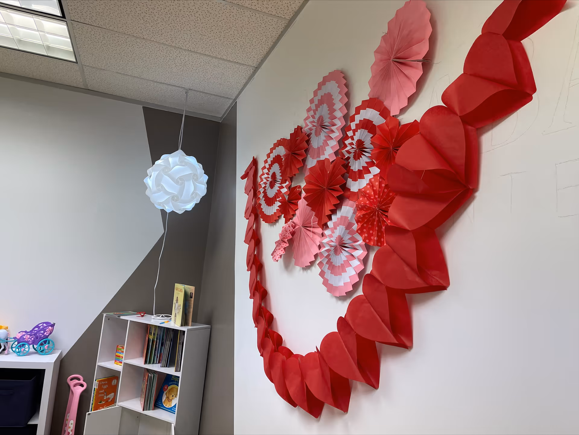 Interior playroom with red paper hearts garland, white pendant light, bookshelf and toys.