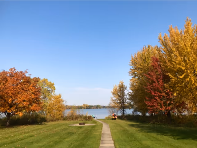 Paved path through a grassy park with autumn trees by a lake.