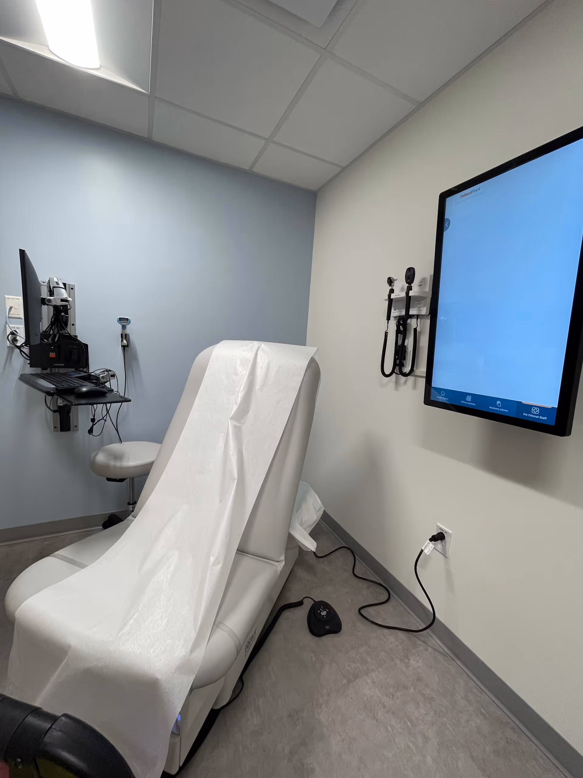 White examination chair in a clinical room with a wall-mounted monitor.