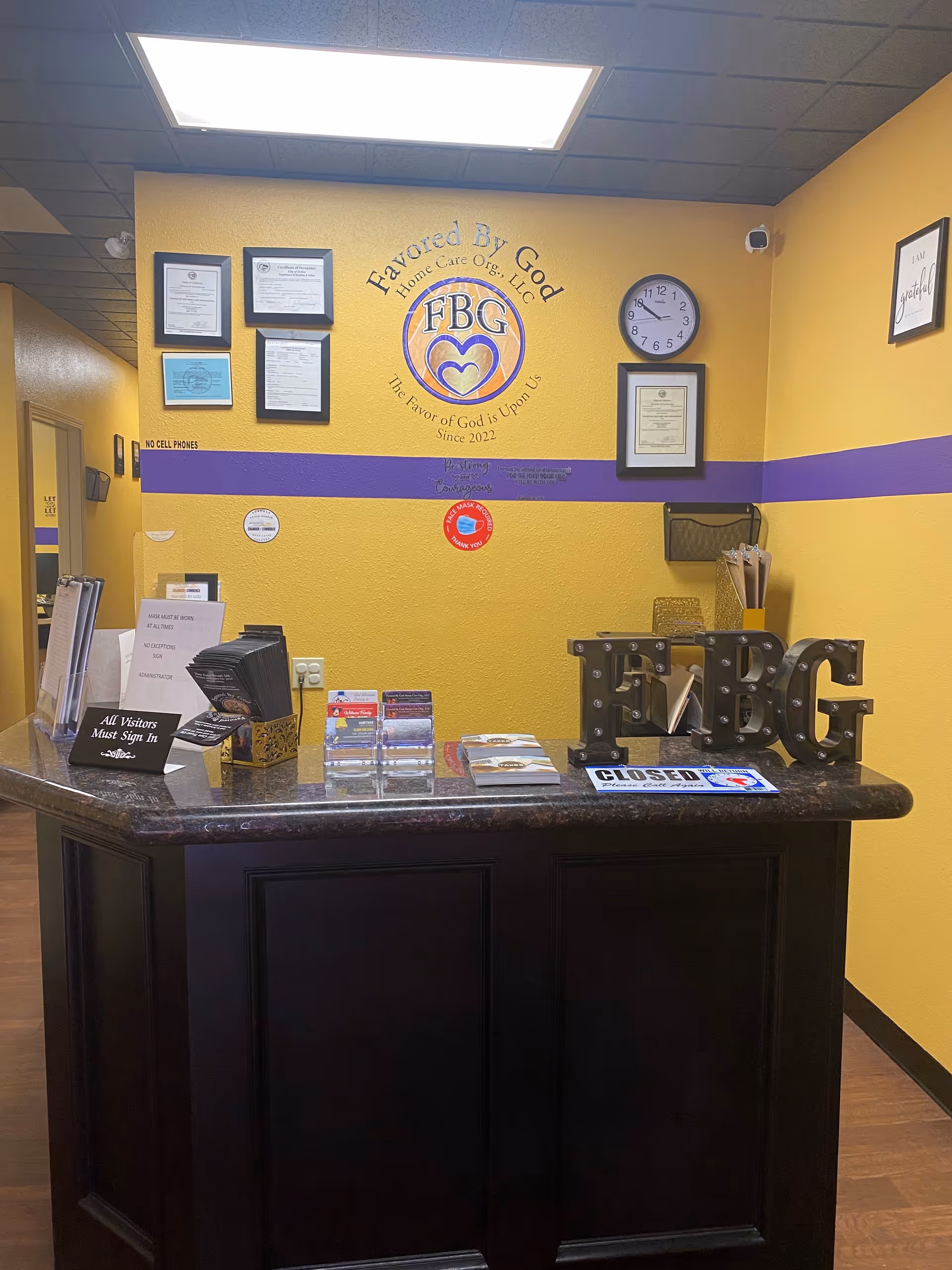 Reception desk in a yellow-walled lobby with certificates, clock, and FBG logo