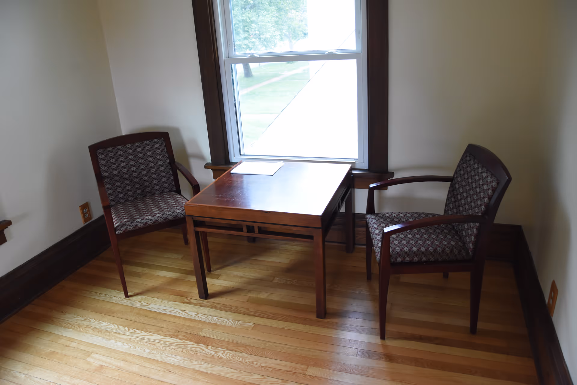 Two patterned chairs sit beside a wooden table by a window in a small interior room.