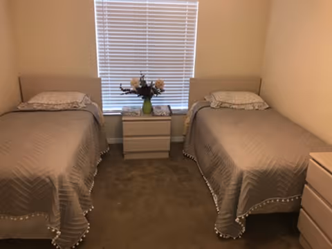 Two single beds with matching gray quilts, a central nightstand, vase, and window blinds.
