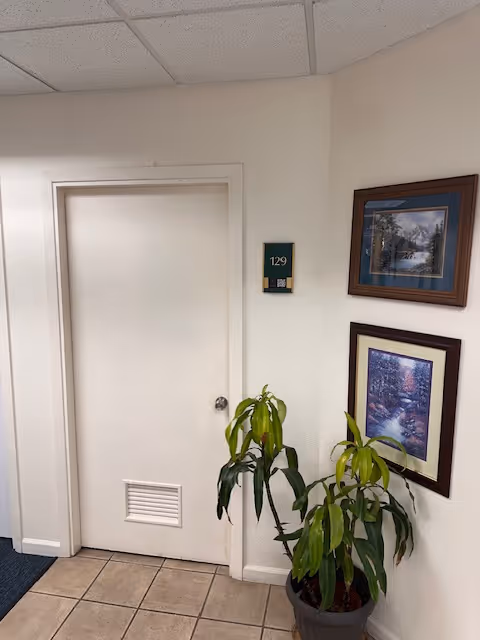 Interior hallway with a closed white door, framed art, and a potted plant.