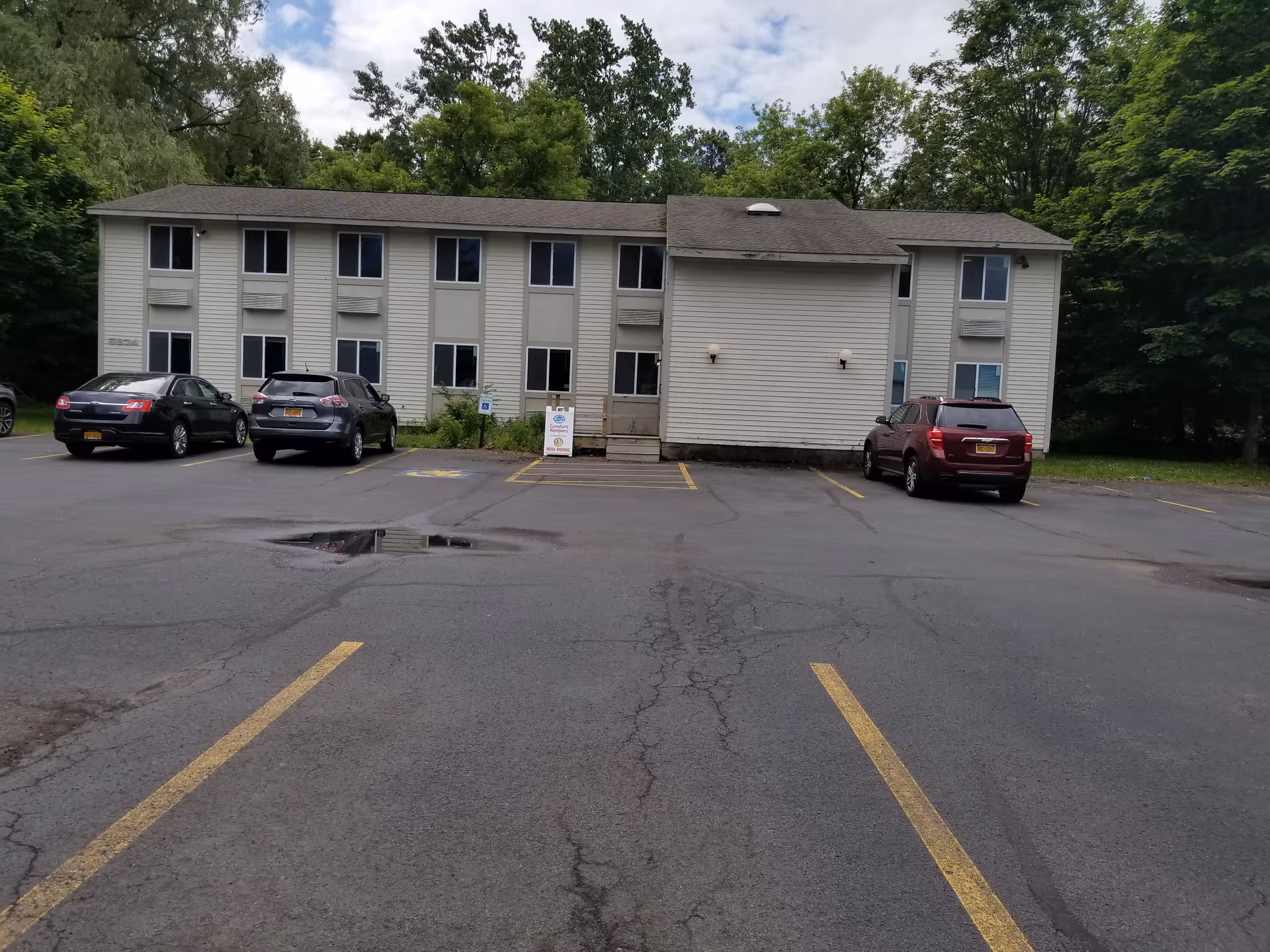 Two-story beige senior living building with cars parked in front and surrounding trees.