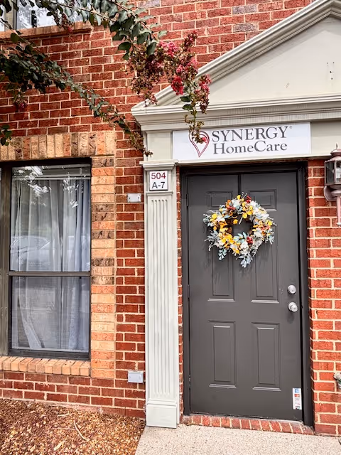 Brick building entrance with a dark door, wreath, window, and Synergy HomeCare sign.