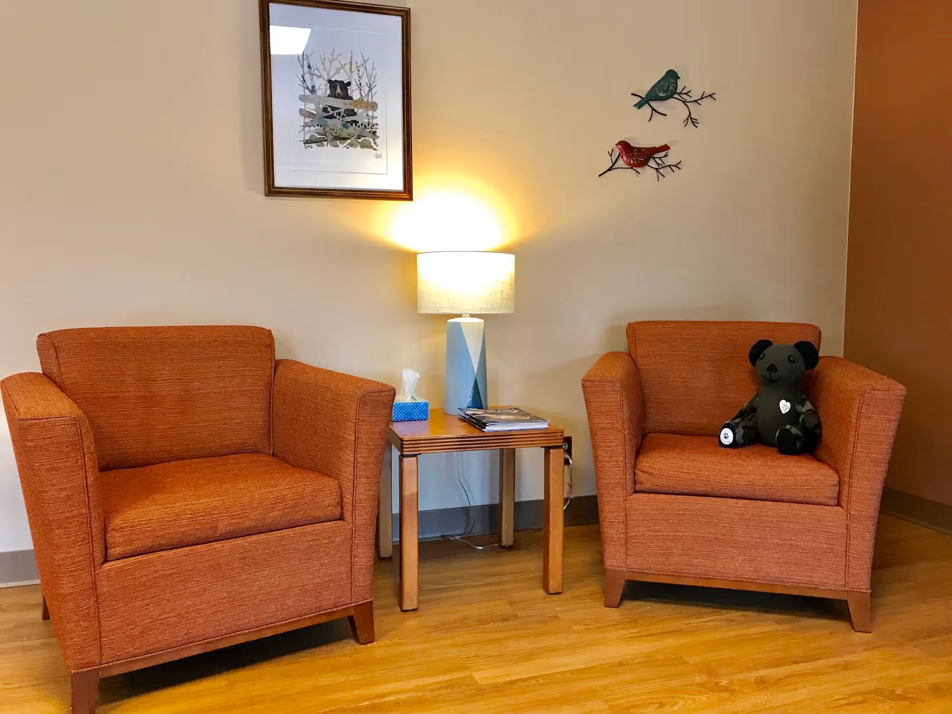Two orange armchairs flank a wooden side table with a lamp in a cozy waiting area.