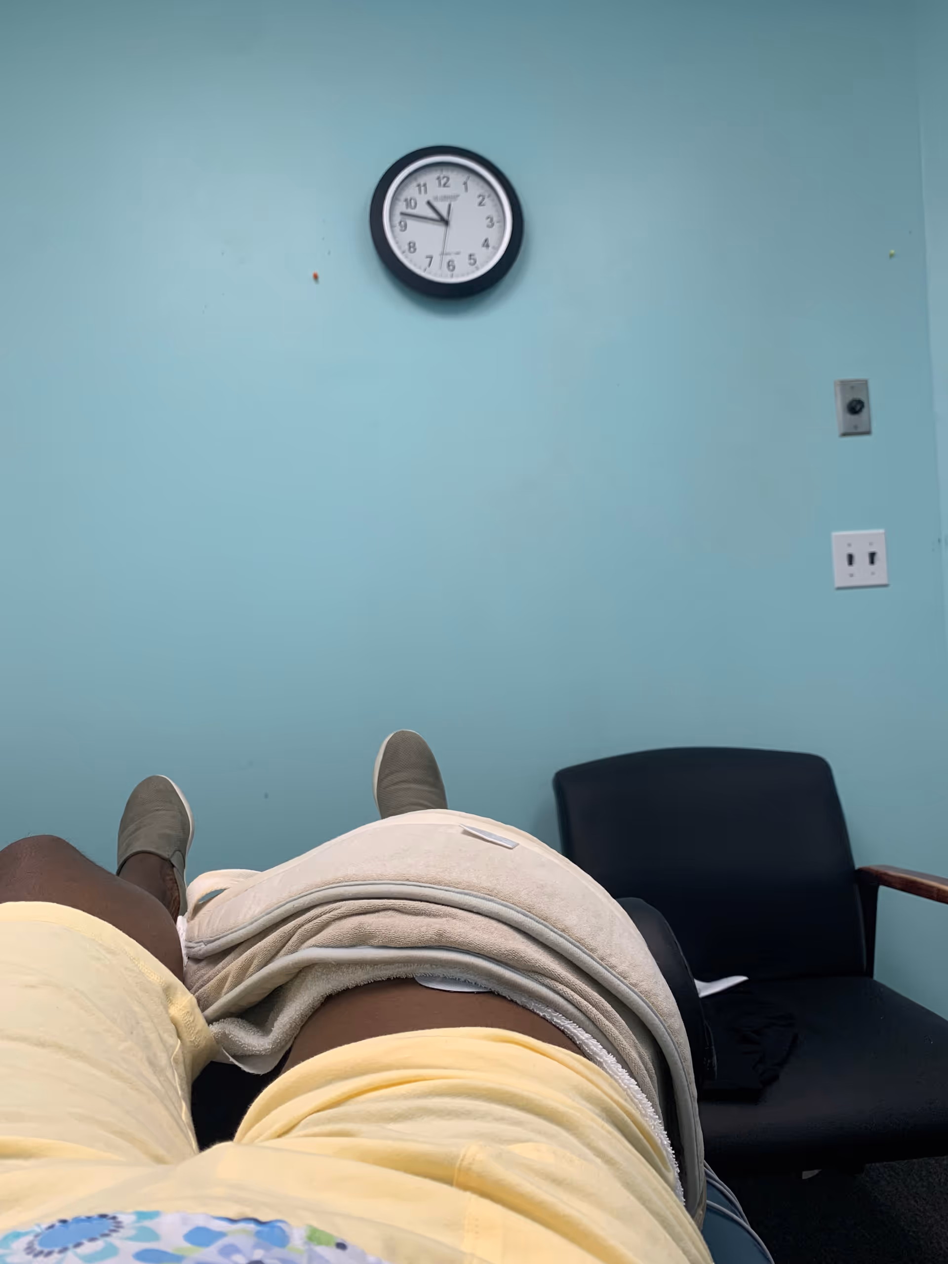 Patient rests on a therapy table in a teal room with a wall clock.