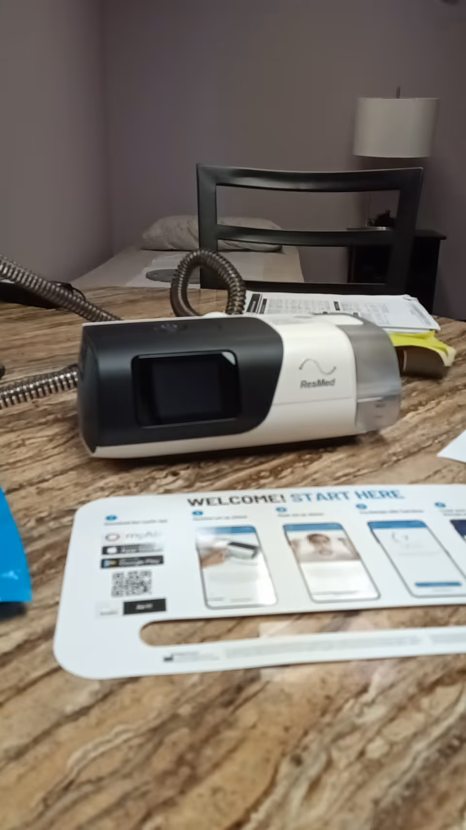 CPAP machine rests on a wood desk with a bed and lamp visible in a bedroom.