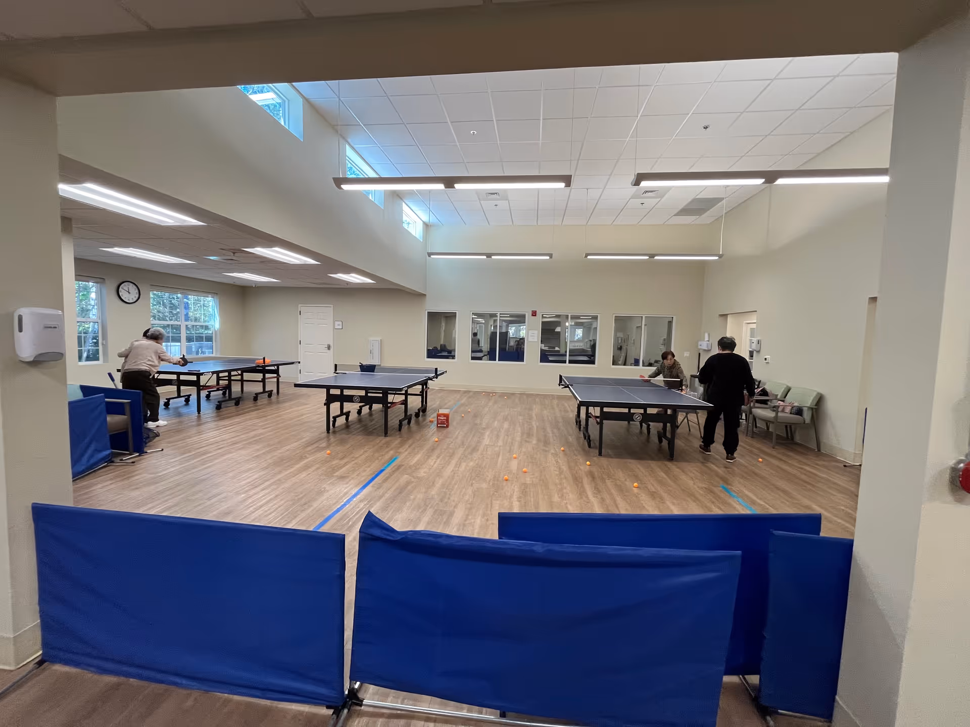 Indoor recreation room with multiple ping-pong tables, players, and blue protective barriers.