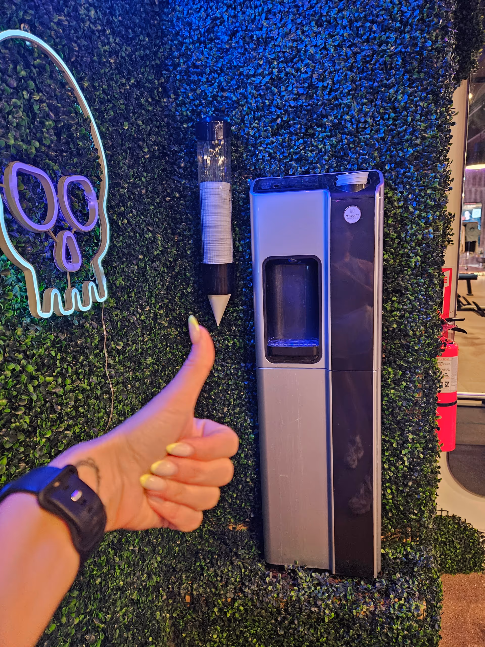 Thumbs-up hand foreground beside a water dispenser mounted on a faux green hedge wall.