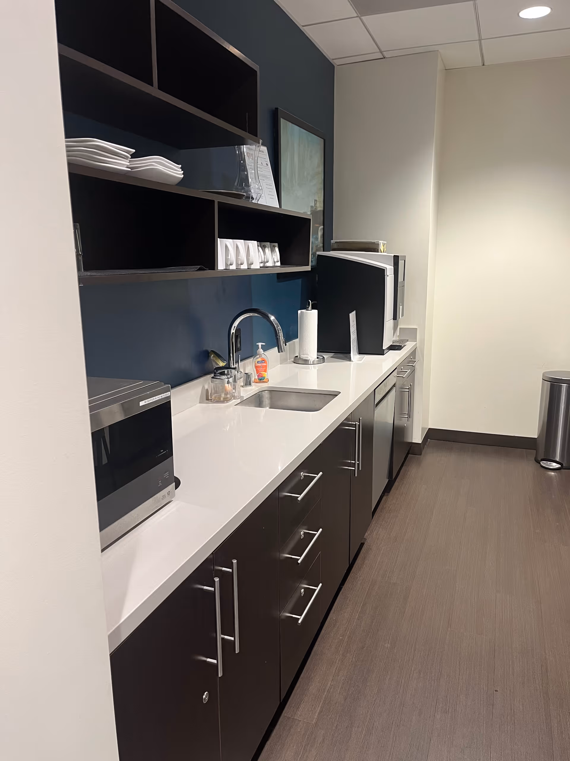 Modern kitchenette with white counter, sink, microwave, dark cabinets, and stacked dishware.