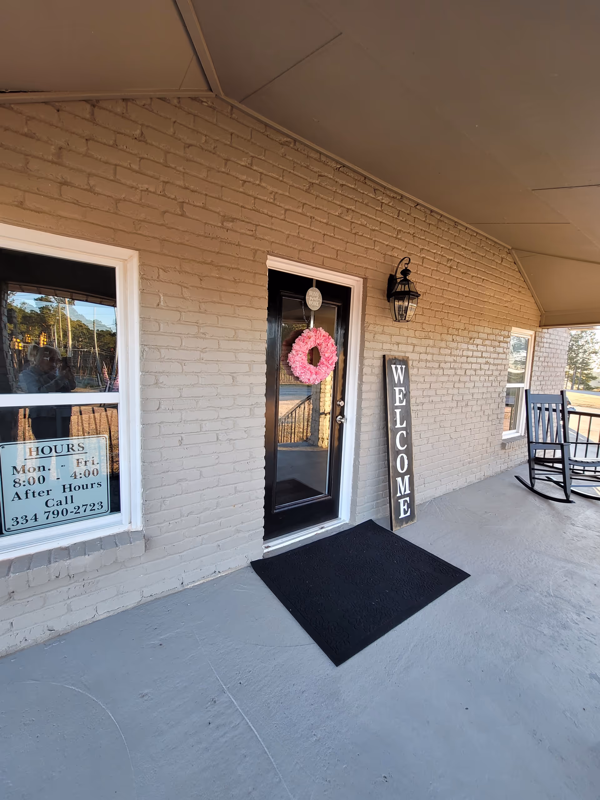 Exterior of a facility entrance with a wreath, welcome sign, and rocking chairs.