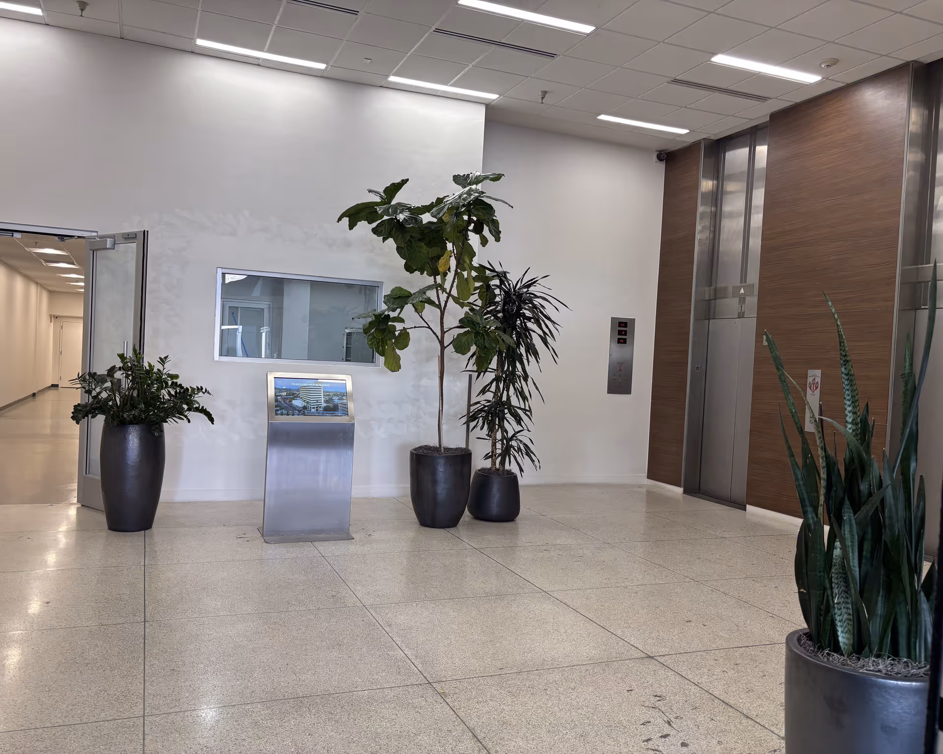 Bright facility lobby with large potted plants, elevator, and a reception kiosk.