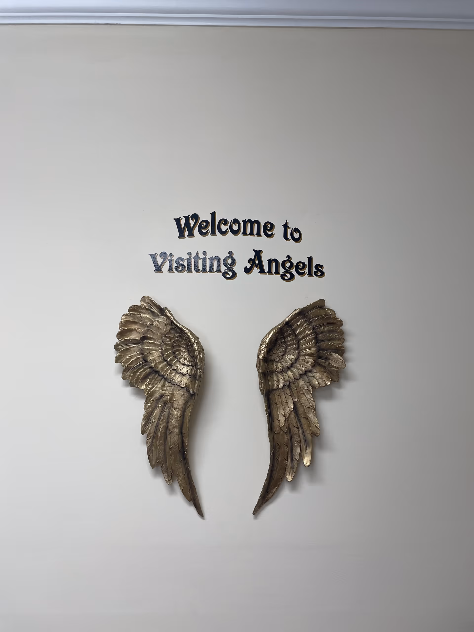Interior wall with decorative bronze angel wings and 'Welcome to Visiting Angels' sign.