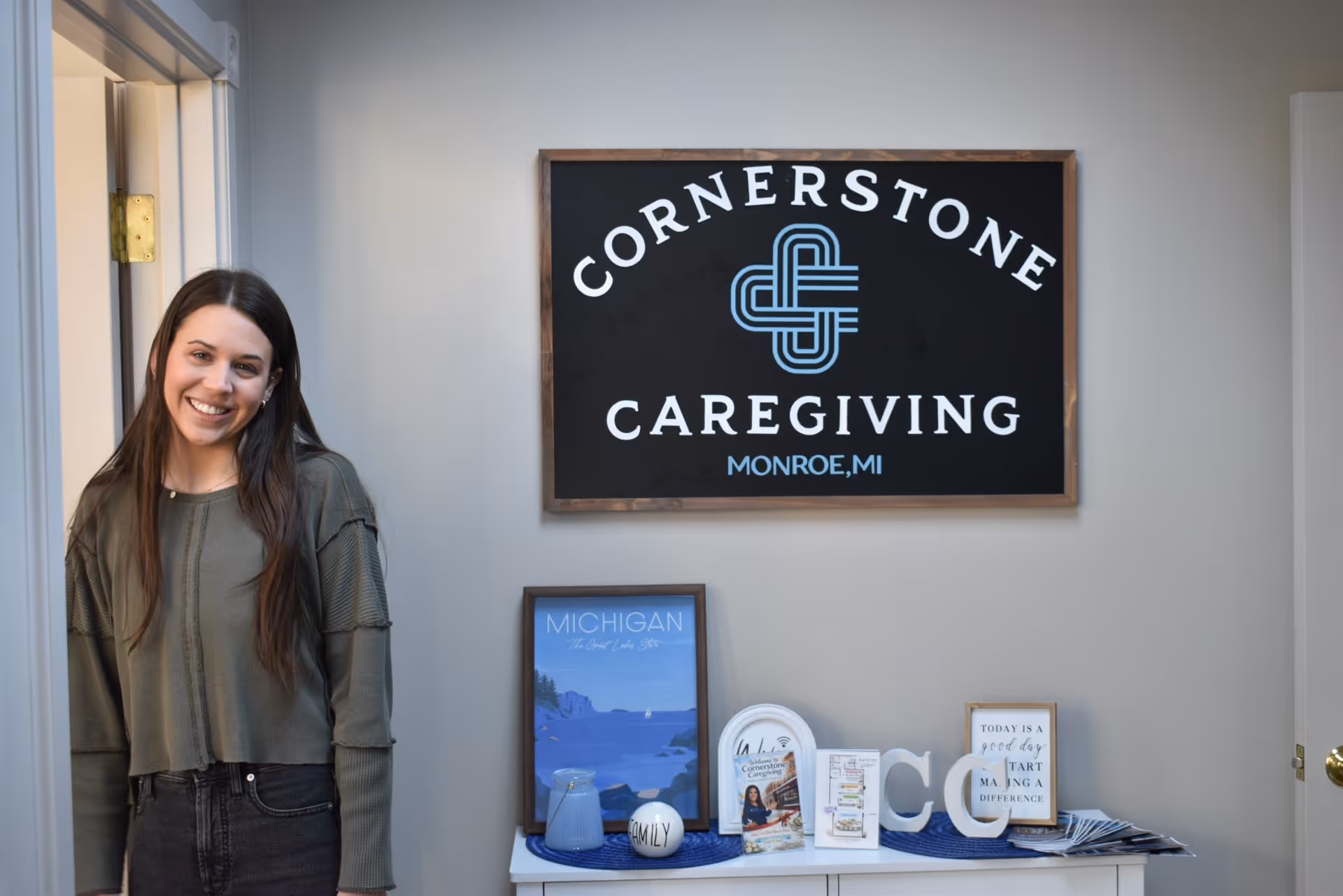 Smiling woman stands beside a Cornerstone Caregiving sign in a welcoming interior lobby, Monroe, MI.