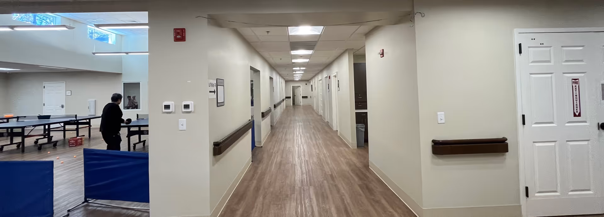 Indoor corridor of a senior living facility with a person near a table tennis setup.