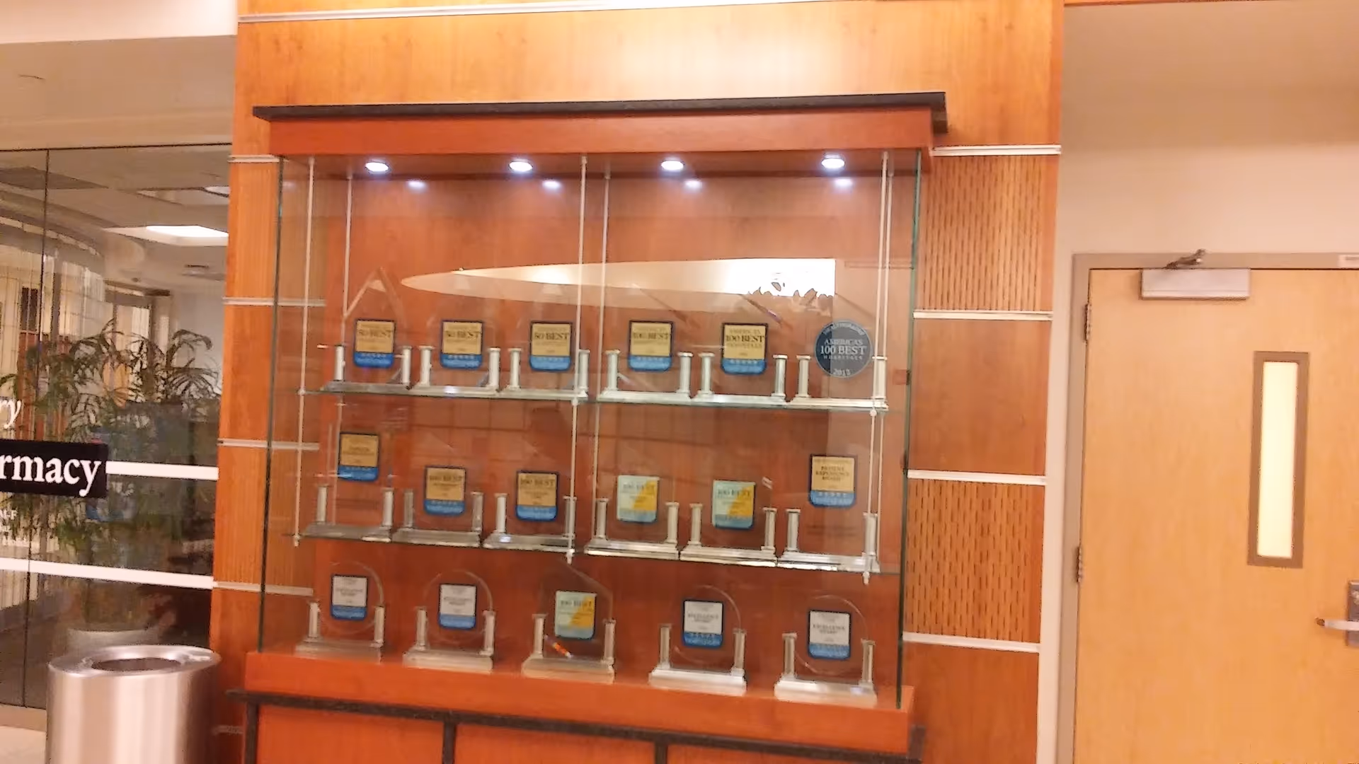Wooden-framed glass display case filled with plaques and awards in a hallway near a door.