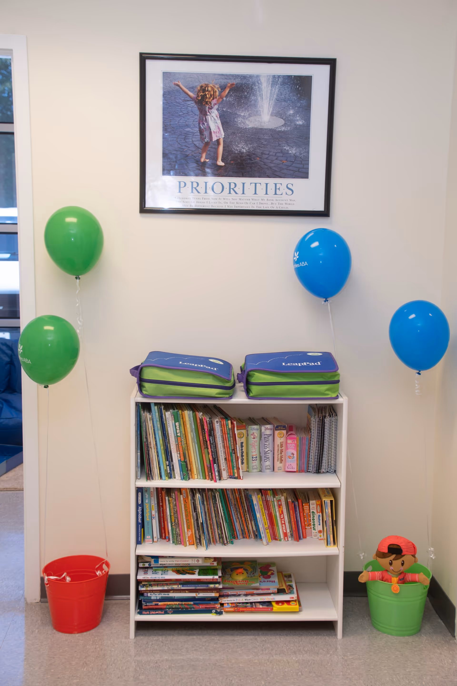 White bookshelf with children's books, backpacks on top, and colorful balloons in a bright interior.