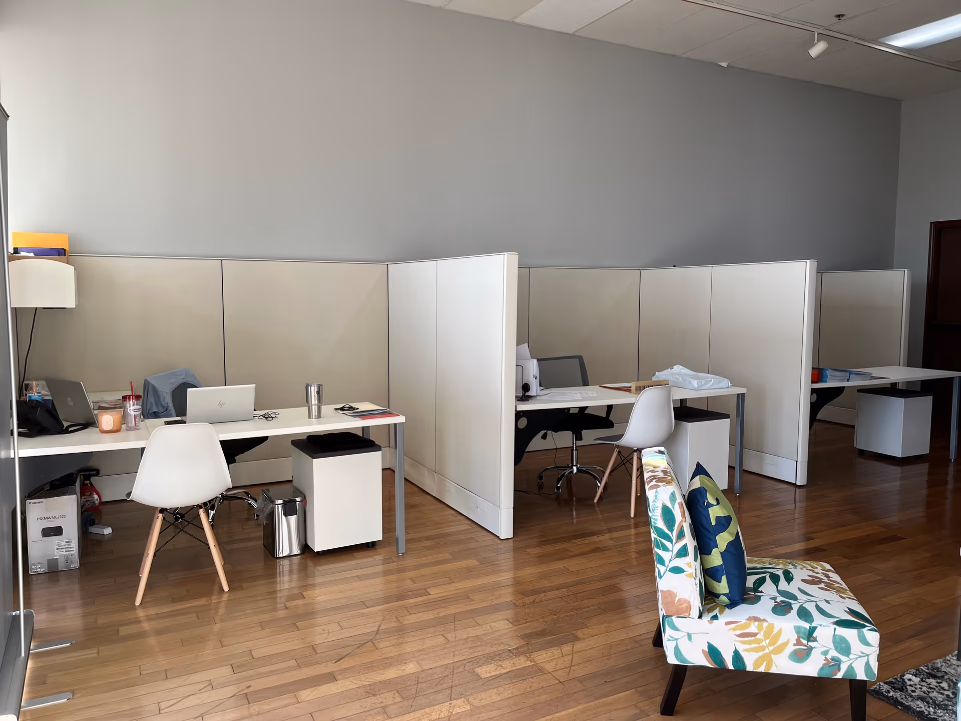 Open-plan office area with cubicles, desks, laptops, and a floral-patterned chair.