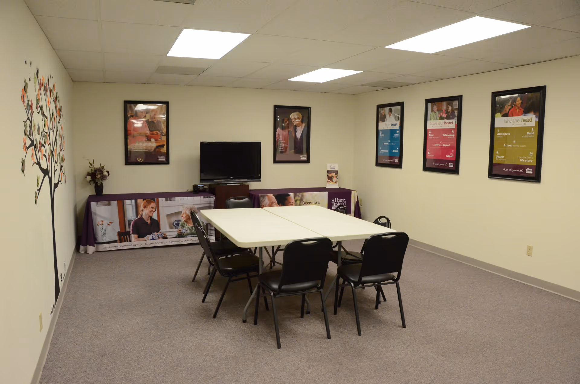 Interior senior living activity room with a central table, chairs, a TV, and posters.
