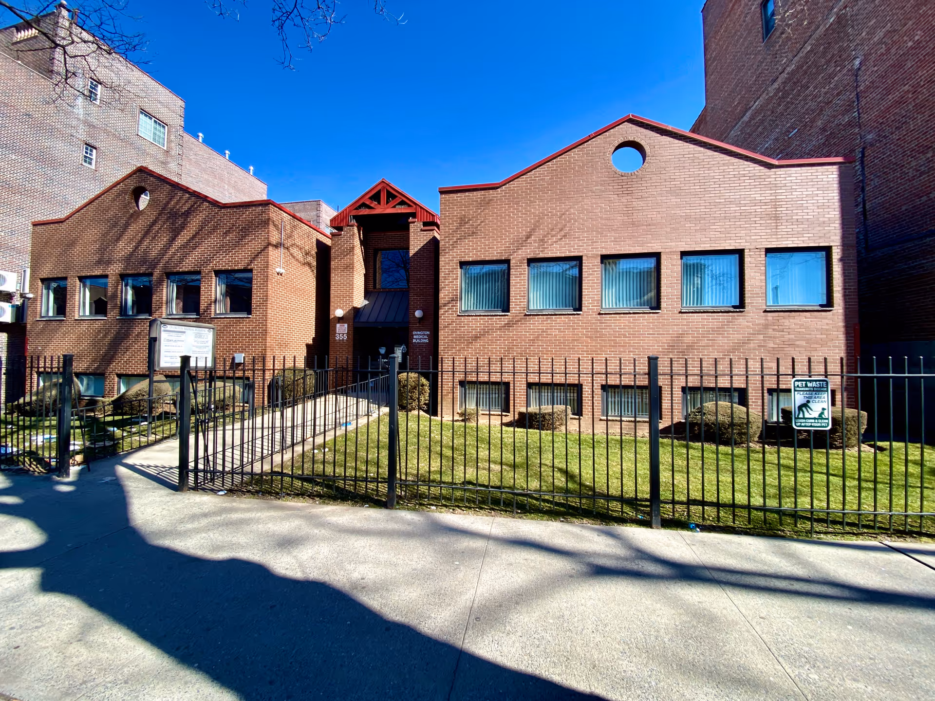 Brick senior living facility exterior with front entrance, metal fence, and blue sky.