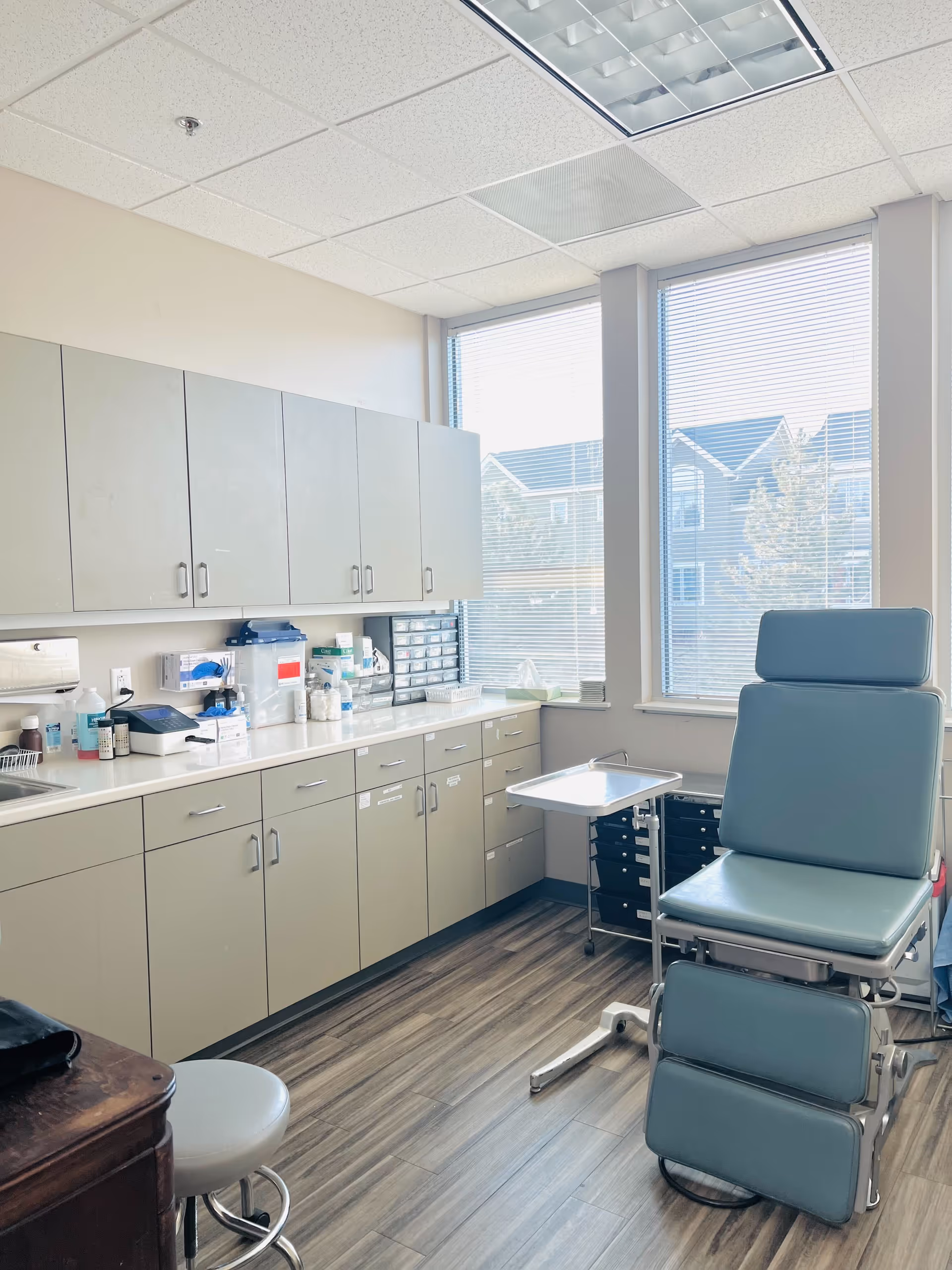 Medical examination room with cabinets, counter, and blue exam chair by windows.