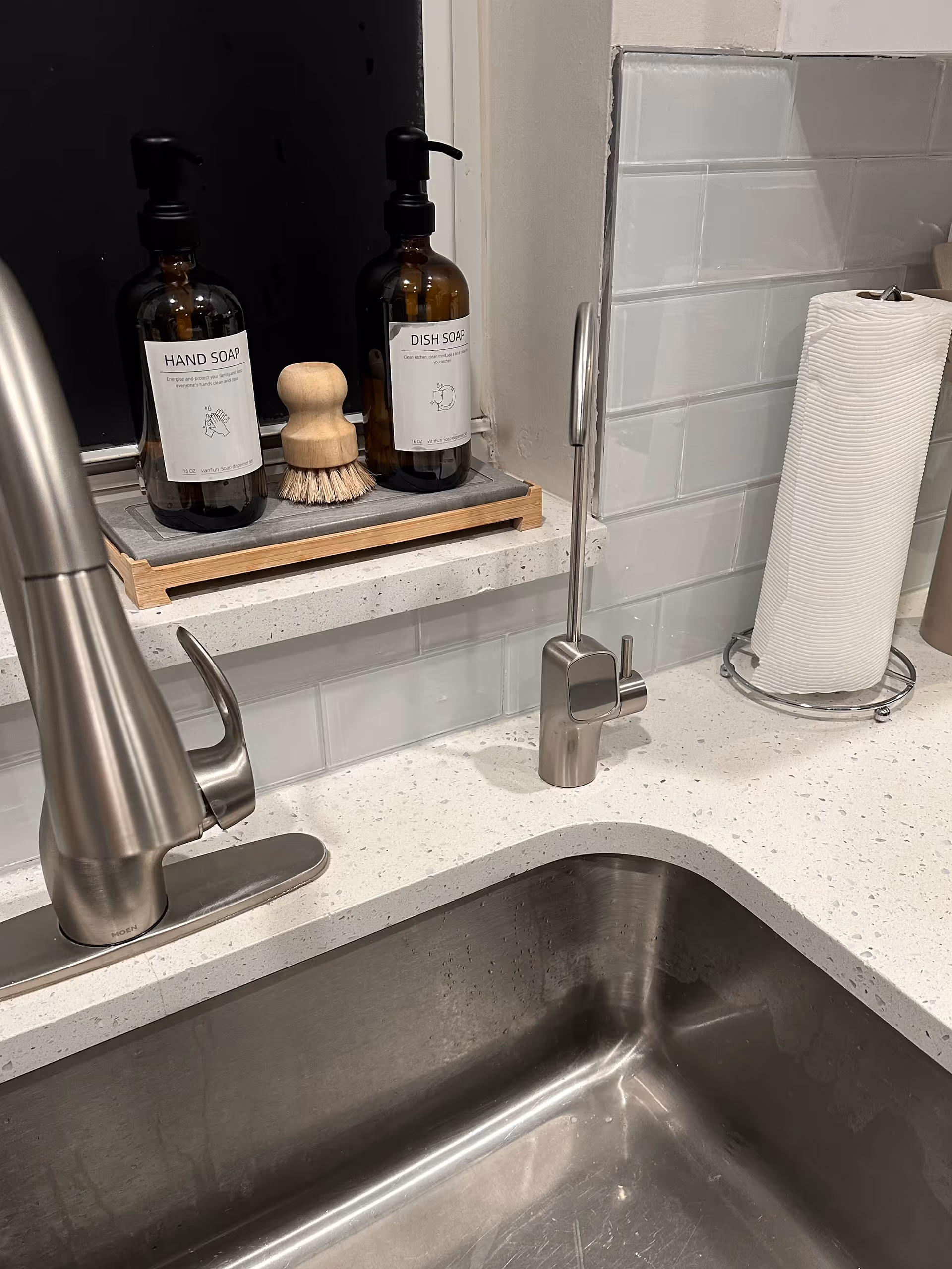 Stainless steel sink and faucet with soap dispensers, brush, and paper towel roll on counter.