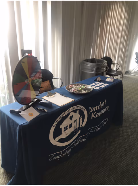 Information table with a colorful wheel, brochures, and Comfort Keepers banner in an indoor space.