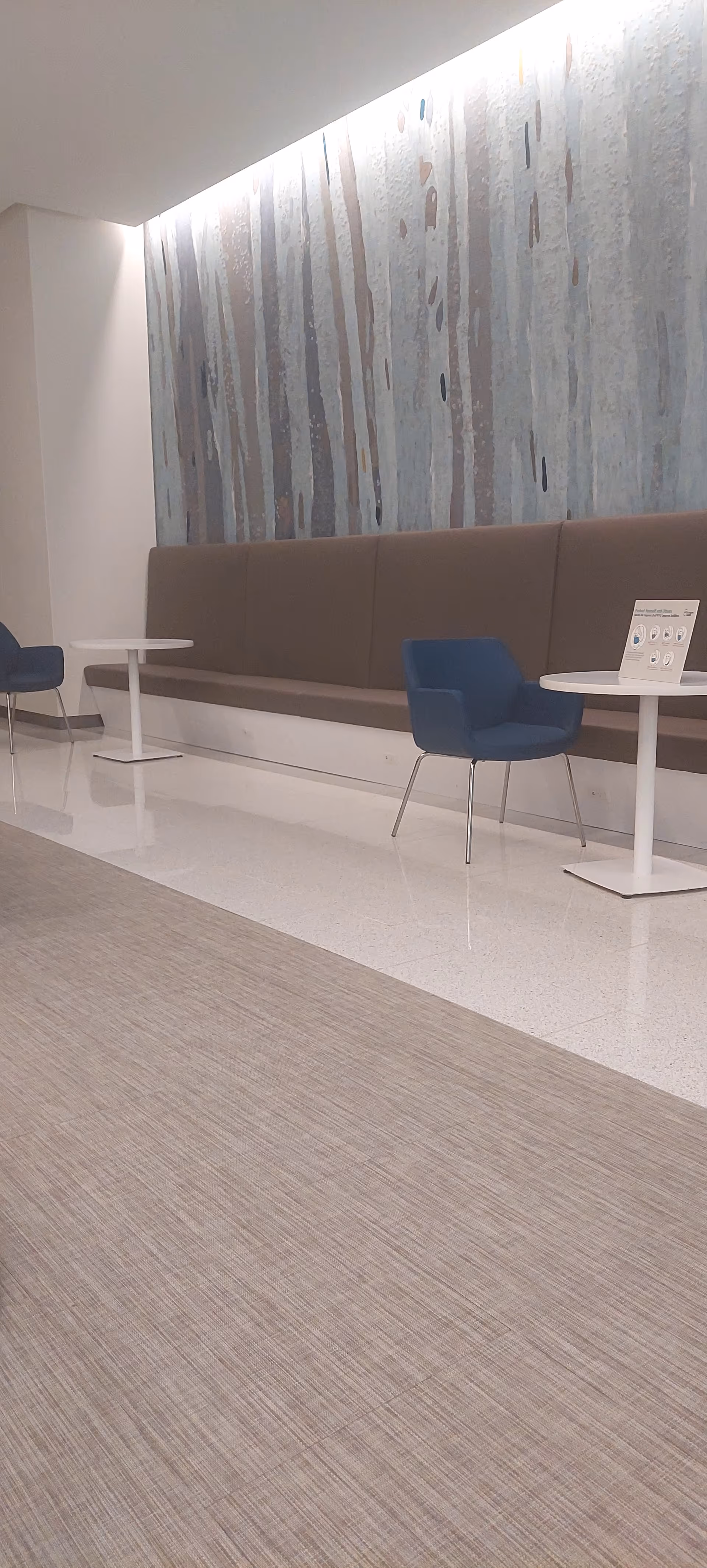 Interior lounge with a long beige sofa, blue chairs, and round white tables.