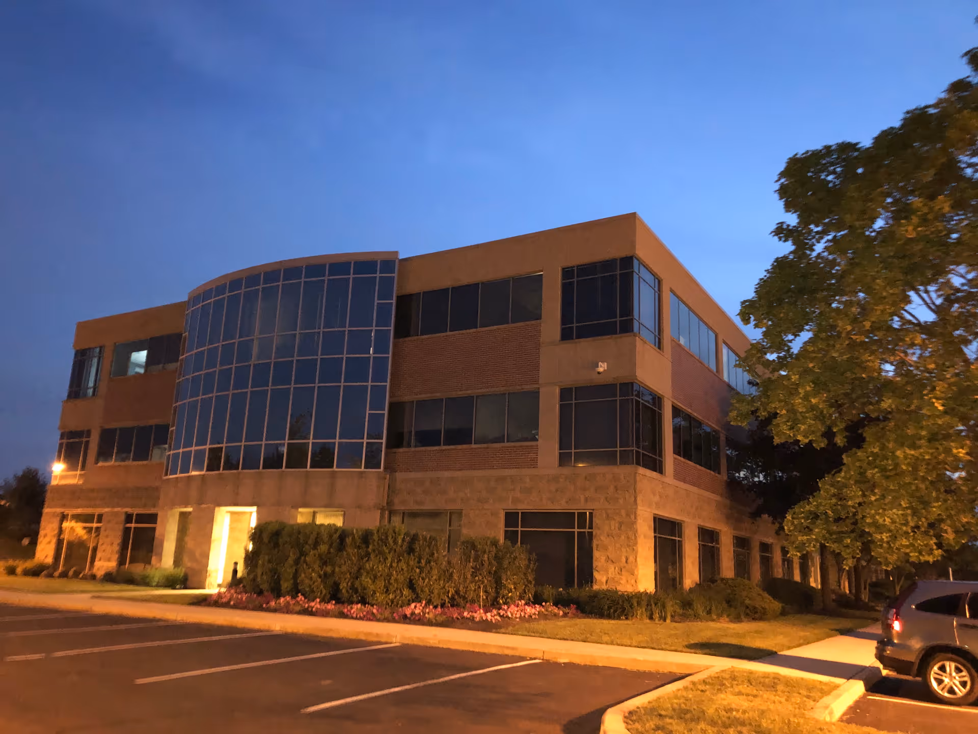 A modern multi-story building exterior at dusk with glass facade and landscaping.