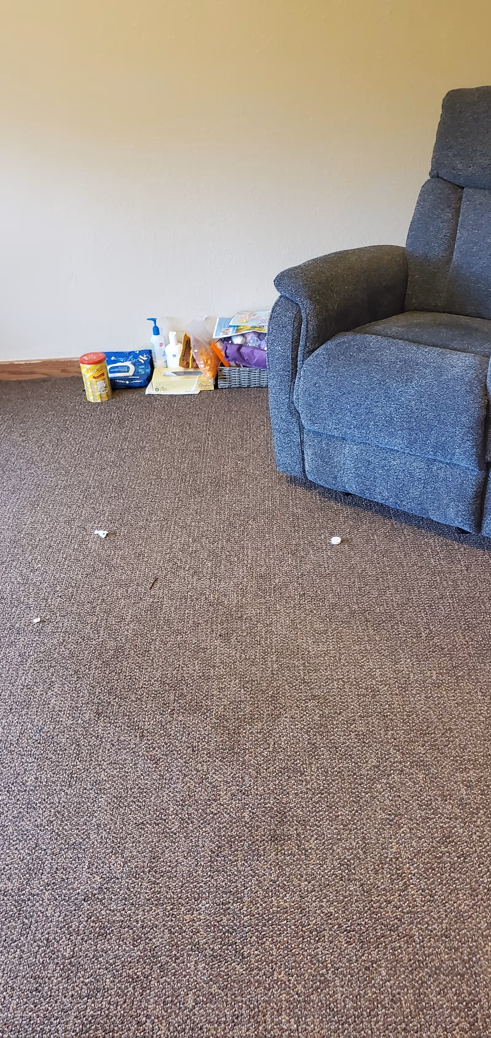 A blue upholstered recliner sits in a living room corner with cluttered items on the floor.
