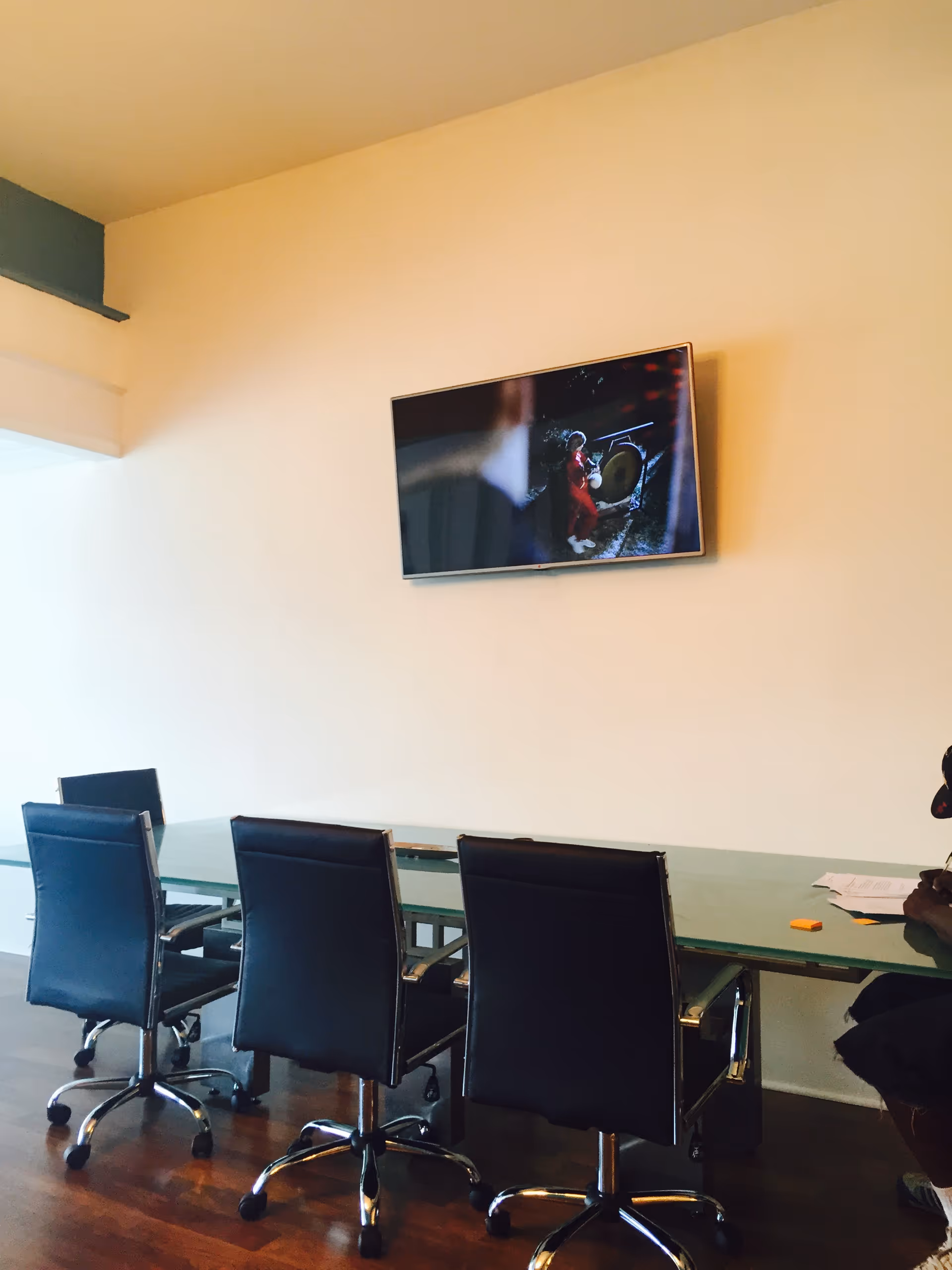 A glass conference table with black rolling chairs and a wall-mounted TV in a bright interior.