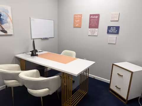 A small office meeting room with a desk, chairs, whiteboard, and wall art.
