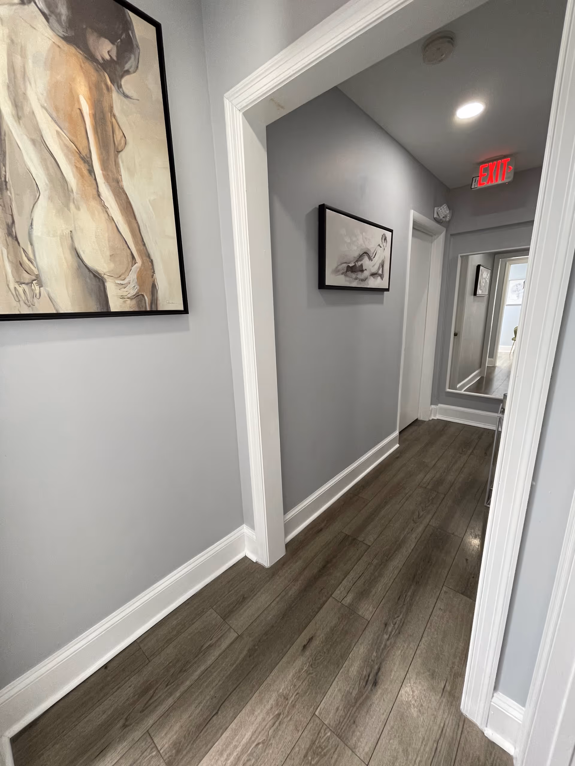 A narrow hallway inside a facility with gray walls, framed art, white trim, and exit sign.