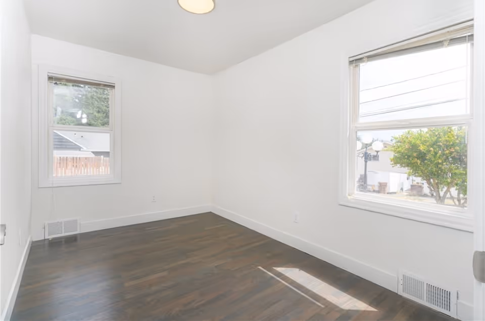 Bright, empty white room with two windows and dark wood flooring.