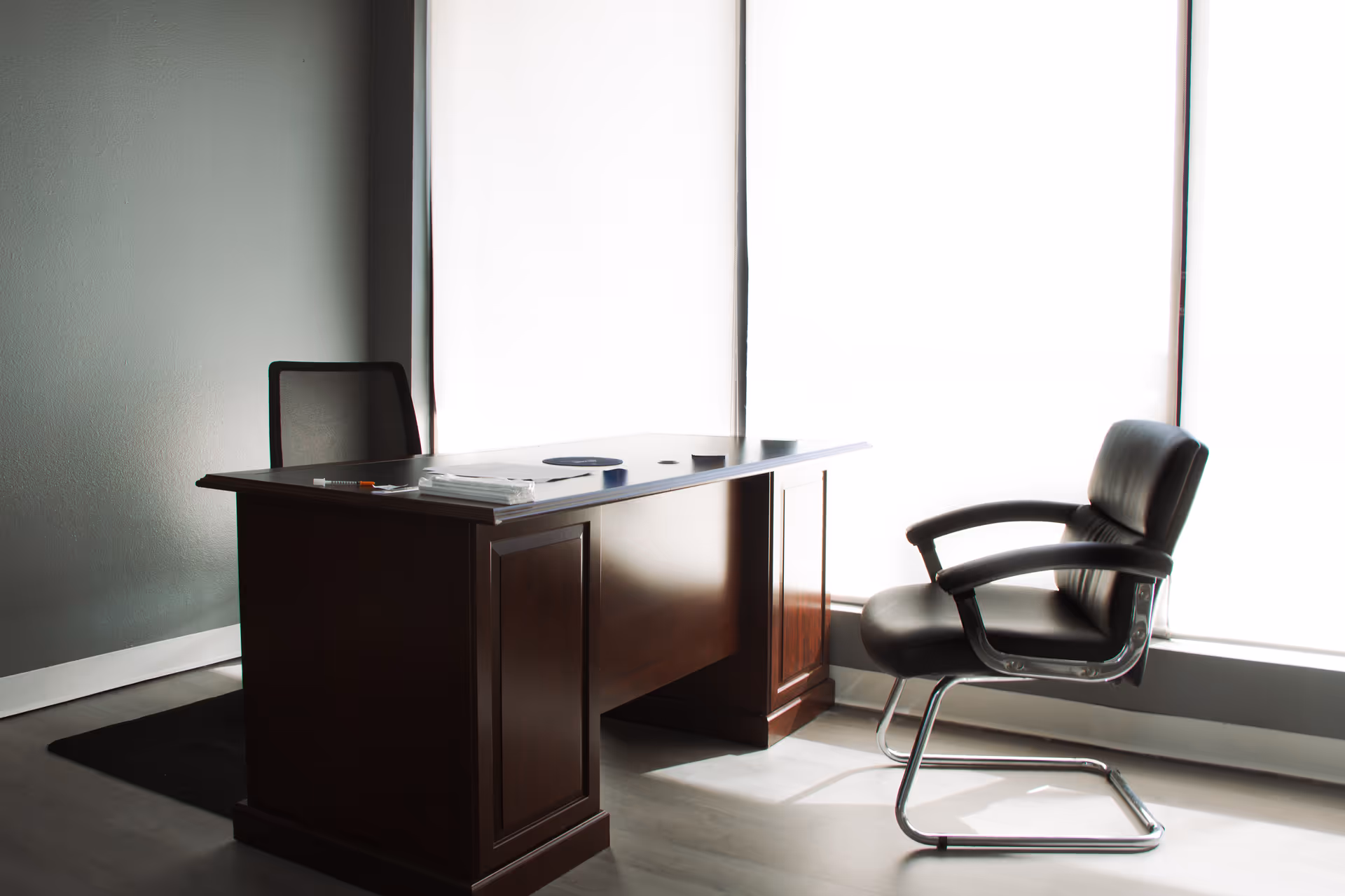 A dark wooden desk and black leather chair beside large bright windows.