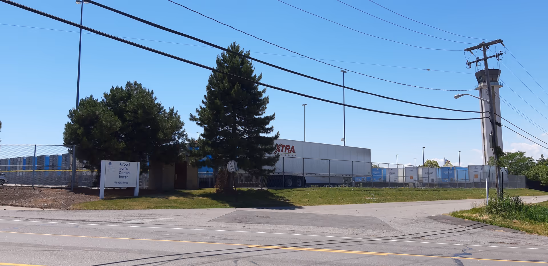 Wide shot of an industrial lot with a chain-link fence, trees, and a white loading trailer.