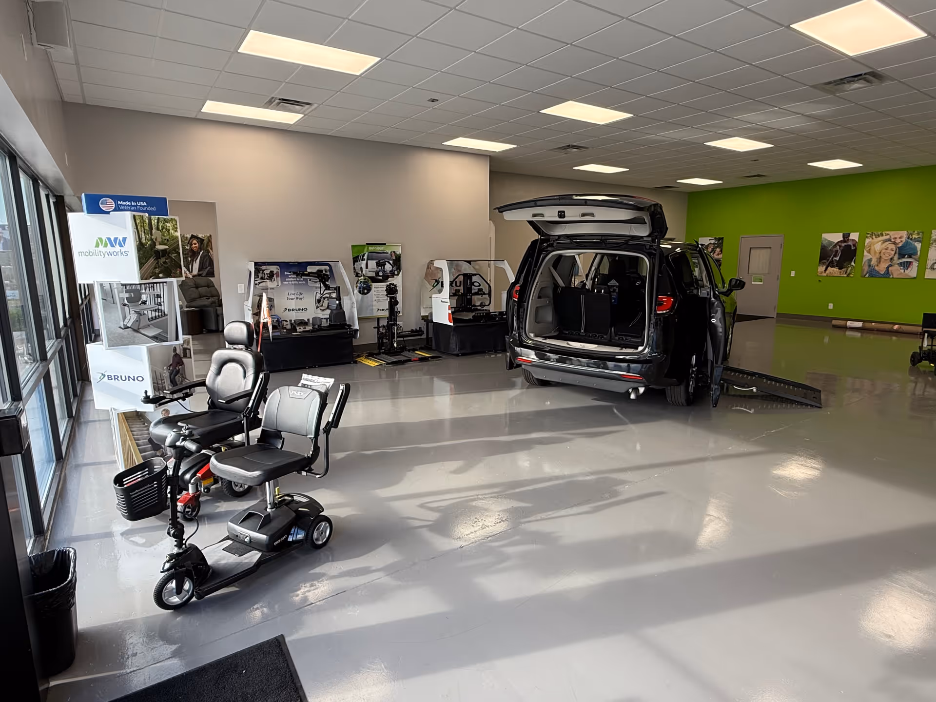 Indoor mobility showroom with a ramp-equipped van, scooters, and display stands along a bright green wall.