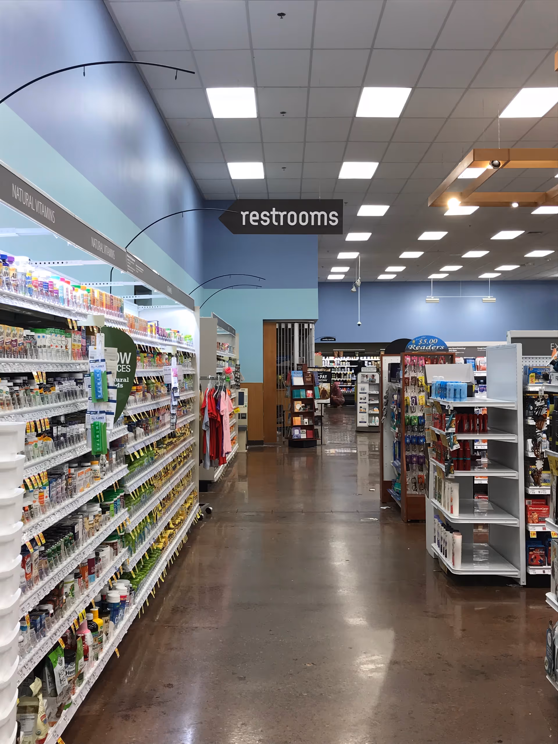 Inside Kroger Pharmacy, an aisle of shelves with medications and a restrooms sign overhead.