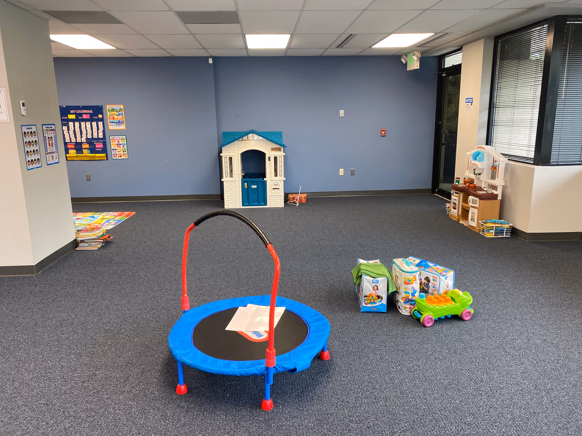 Indoor playroom with a blue-trimmed trampoline, toy house, and scattered children's toys.