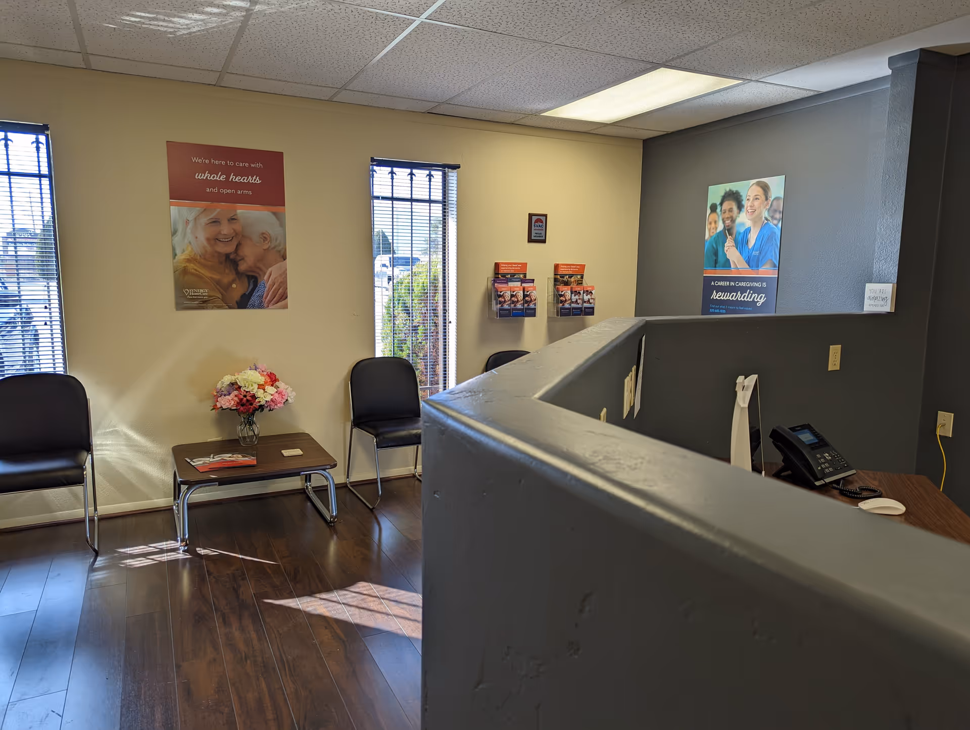 Reception area with chairs, a small table, flowers, posters, and a curved counter.